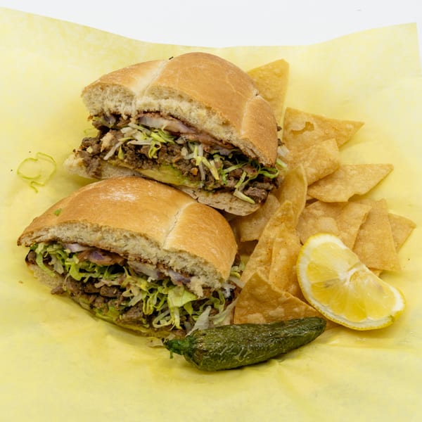 Two halves of a steak torta with lettuce, onions, and avocado, served with tortilla chips, lemon, and a jalapeño.