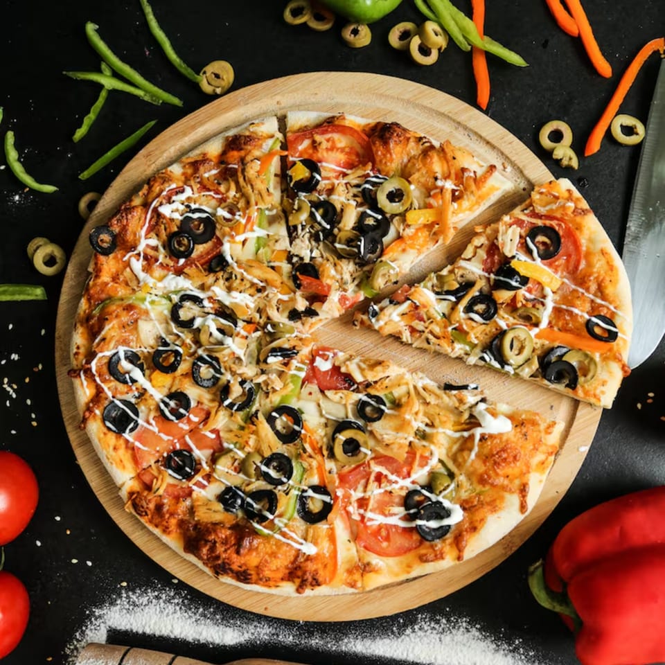 A sliced pizza with olives, chicken, and vegetables on a wooden board, surrounded by ingredients.