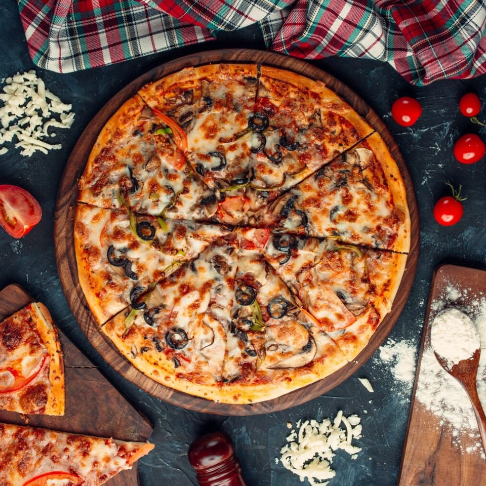 A sliced mushroom and olive pizza on a wooden board, with ingredients and a plaid cloth.