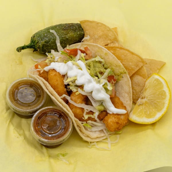 Shrimp tacos with guacamole, sour cream, and shredded lettuce, served with chips, a lemon wedge, and sauces.