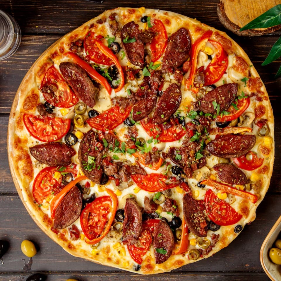 A Neighborhood Pizzeria Y Pollo pizza, topped with pepperoni, tomatoes, olives, and peppers on a wooden table.